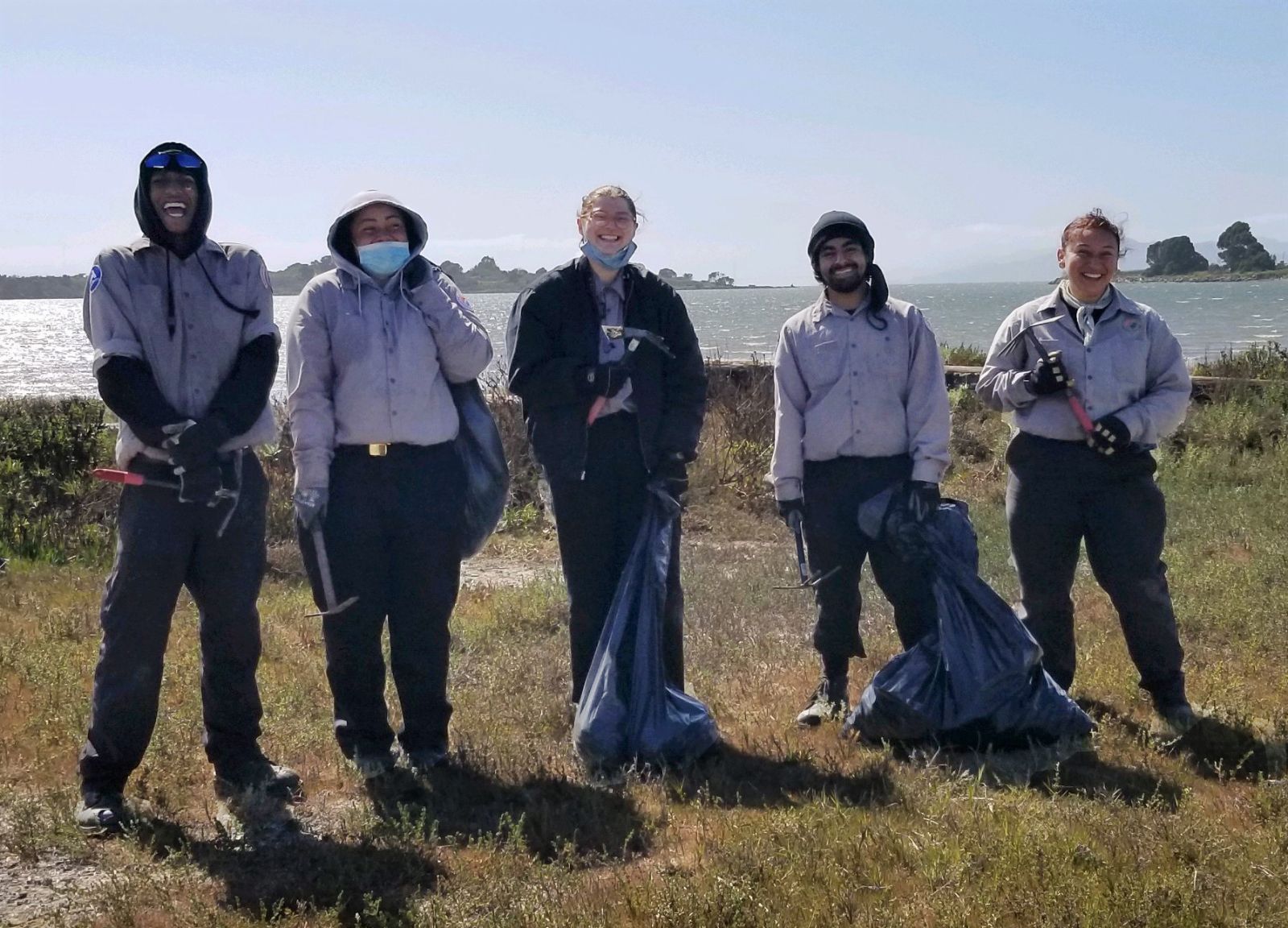 Five Oakland Civicorps members in work clothes stand smiling in a grass field by